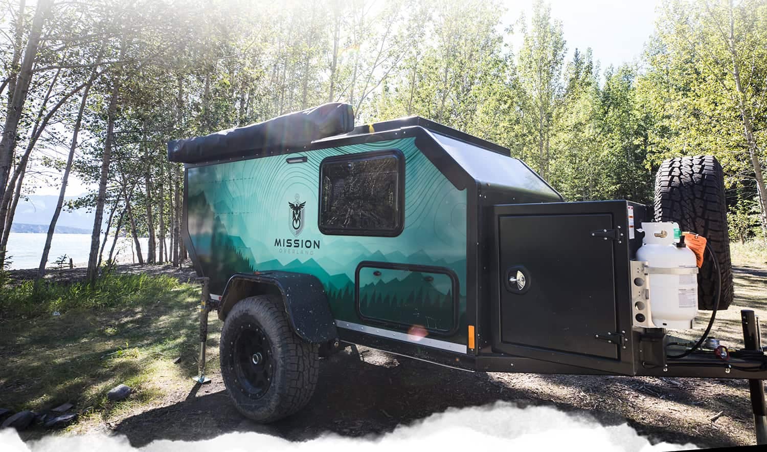 An Mission Overland Trailers camper trailer parked in the woods next to a lake.