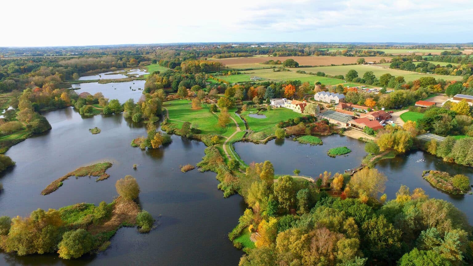 An aerial view of a lake surrounded by lush greenery, with a charming house tucked away amidst the scenery, perfect for eco-tourism enthusiasts seeking a glamping experience at Pensthor