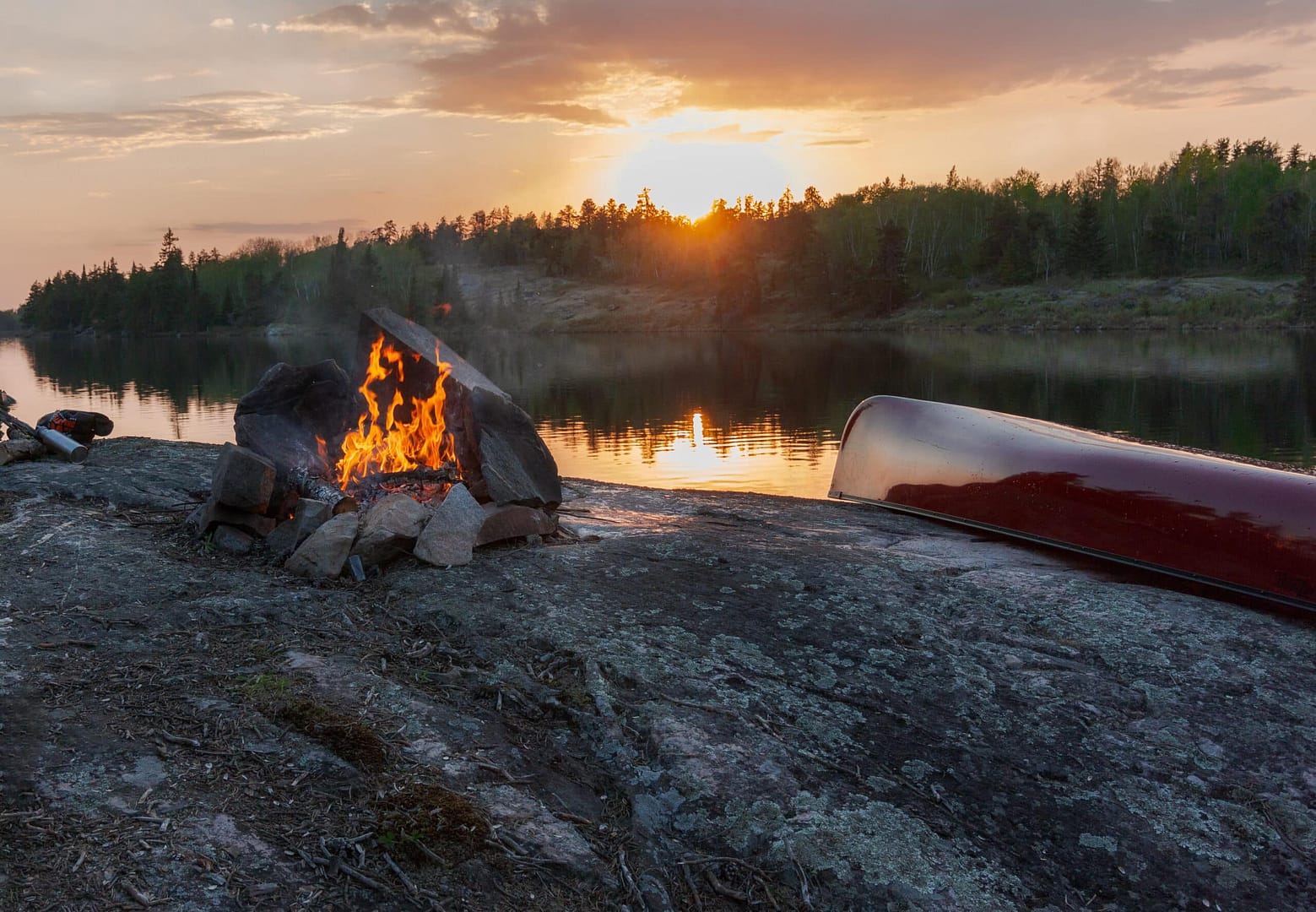 Campfire burning at sunset by a lake with a red canoe overturned on a rocky shore, surrounded by forested hills. It's challenging to extract relevant SEO keywords from this serene scene without additional details.