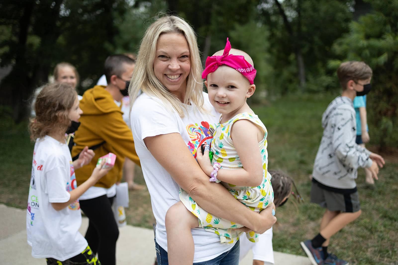 A smiling woman holding a young child in a colorful outfit at an outdoor charity event organized by Care Camps, with other people in the background.
