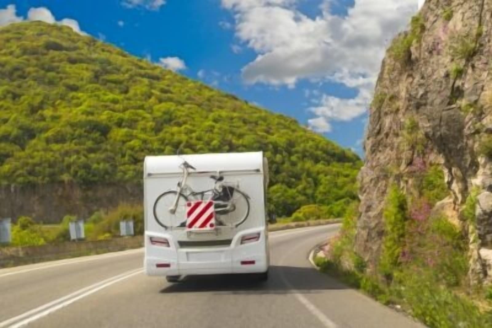 An rv is driving down a mountain road with a bicycle attached to it.