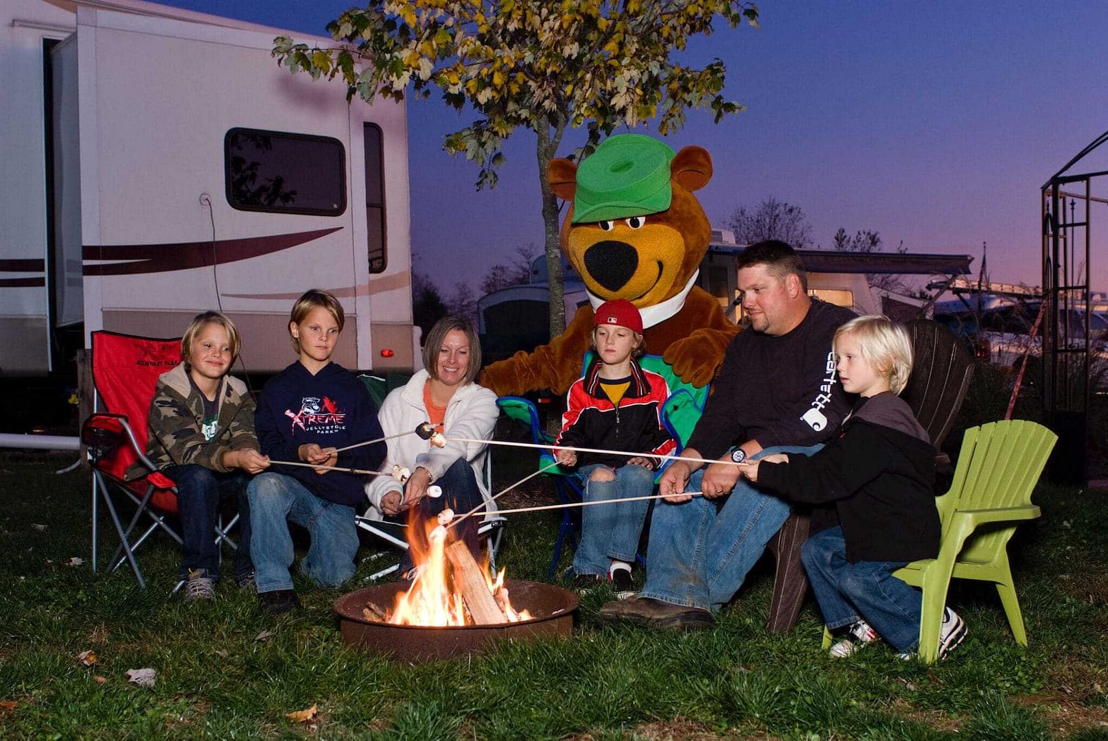 A family sits around a campfire in front of an RV at Jellystone Park.
