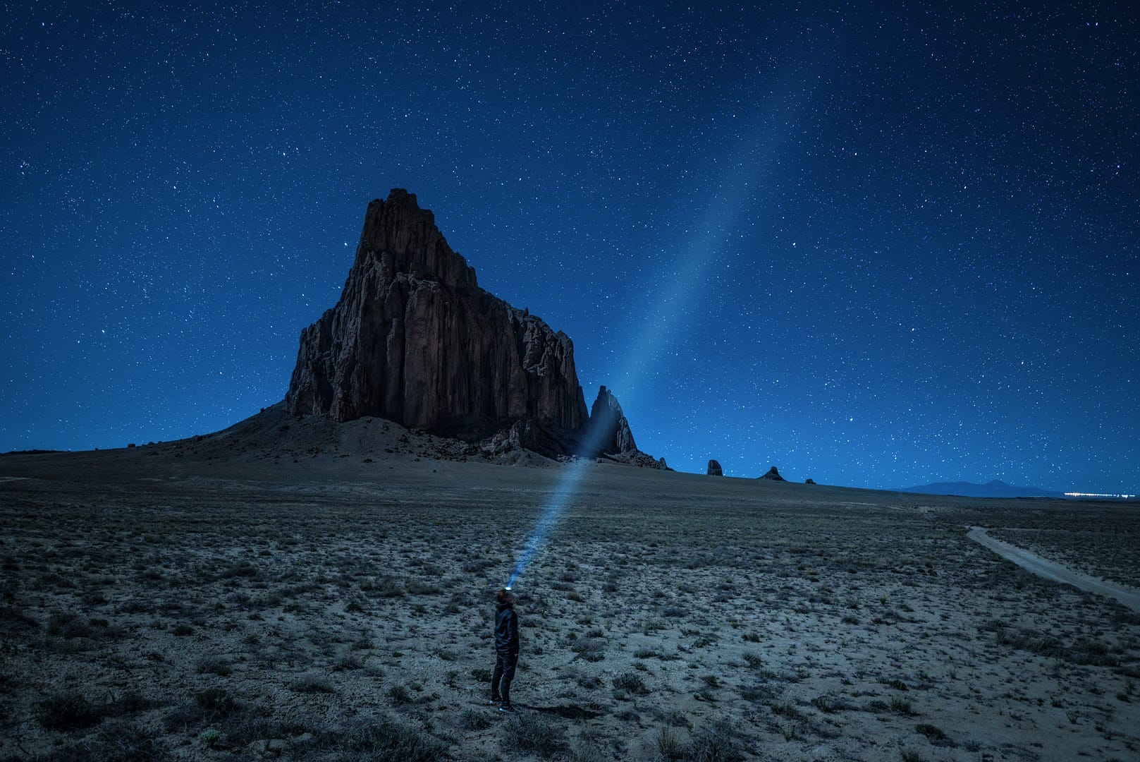 A person stands in the New Mexico desert at night, shining a flashlight beam upwards at a towering rock formation under a starry sky.