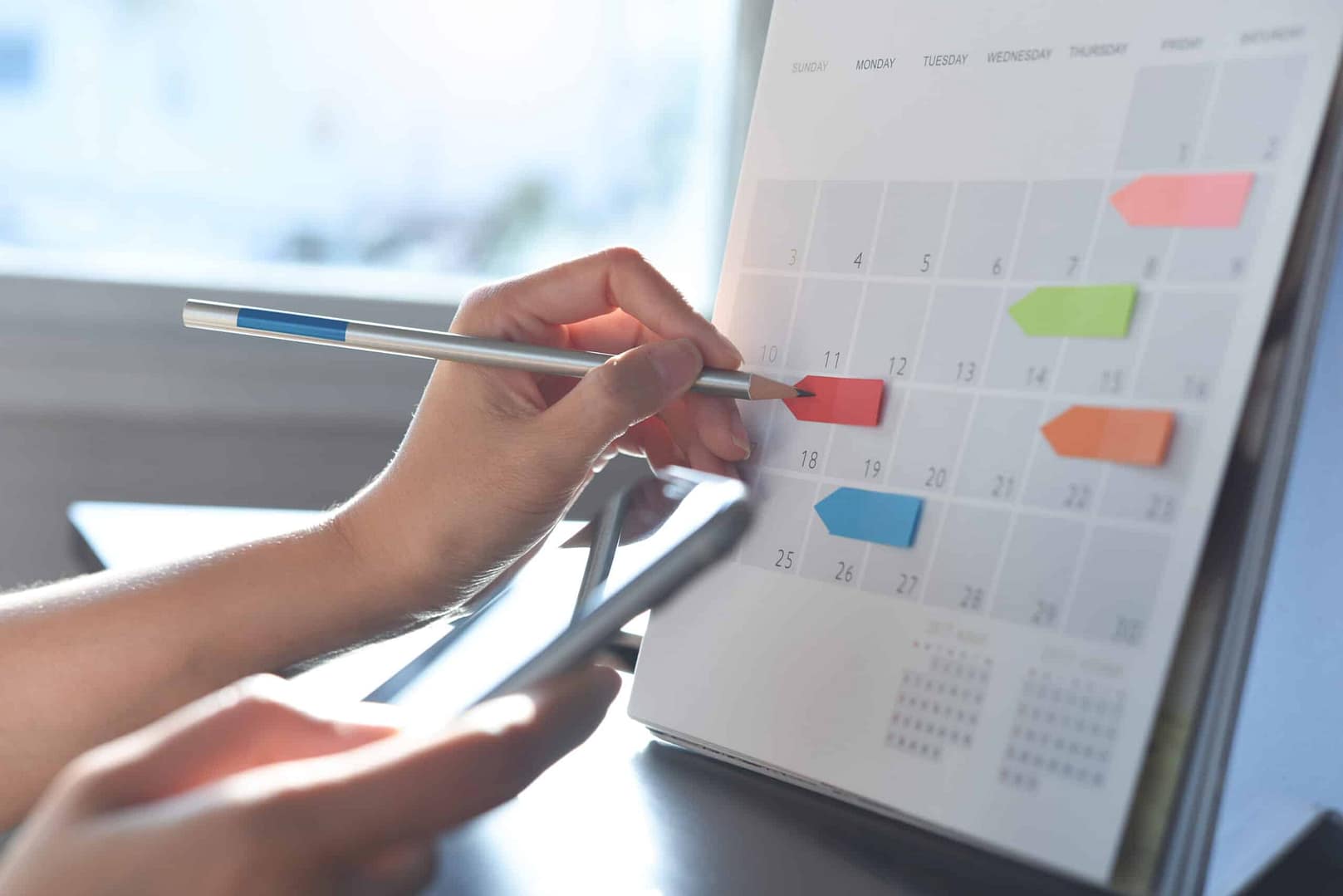 A person using a pen to mark a date on a desk calendar while holding a smartphone in the other hand, planning for festivals and seasonal events. The calendar has colored sticky notes on several dates to attract guests.