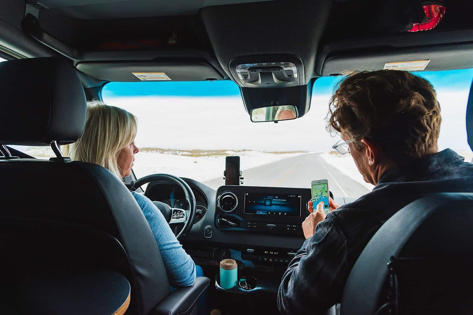 Two diverse people sitting in the back seat of a van looking at their phones.