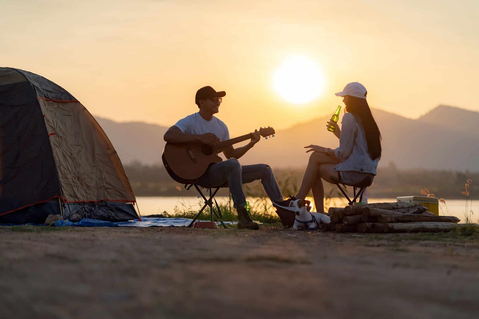 Two people glamping in front of a tent at sunset, playing guitar.