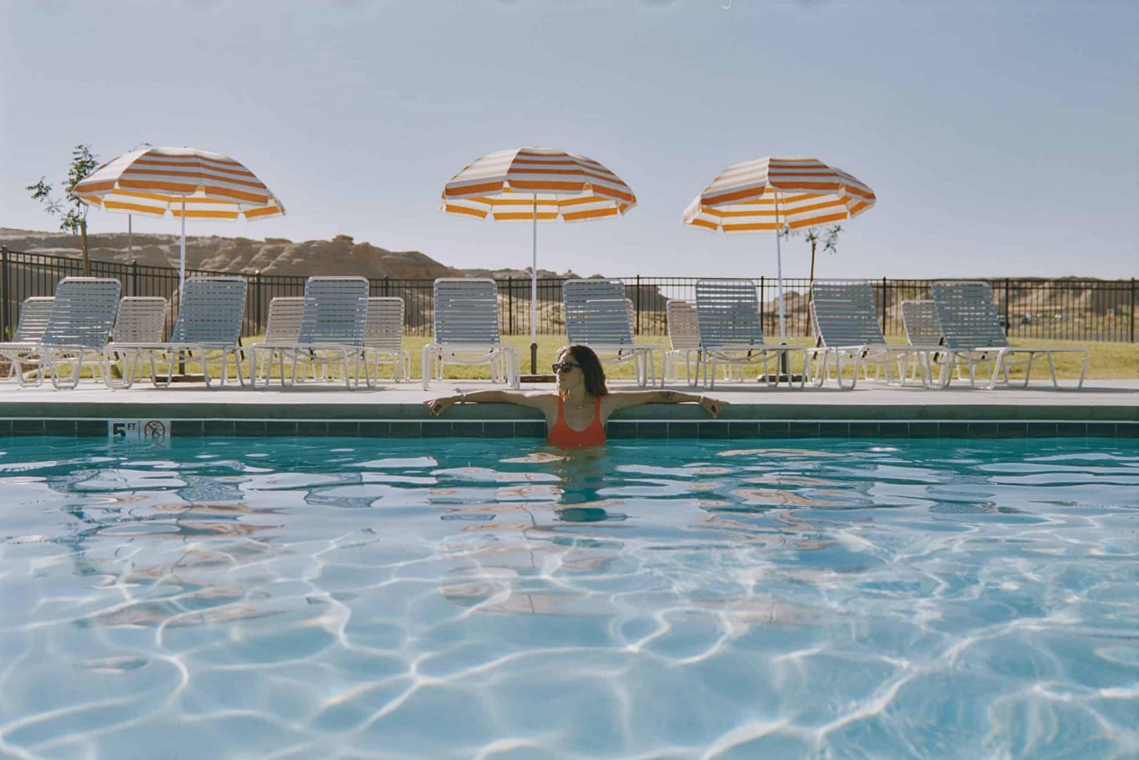 A woman is sitting in a pool with chairs and umbrellas, showcasing modern outdoor hospitality.