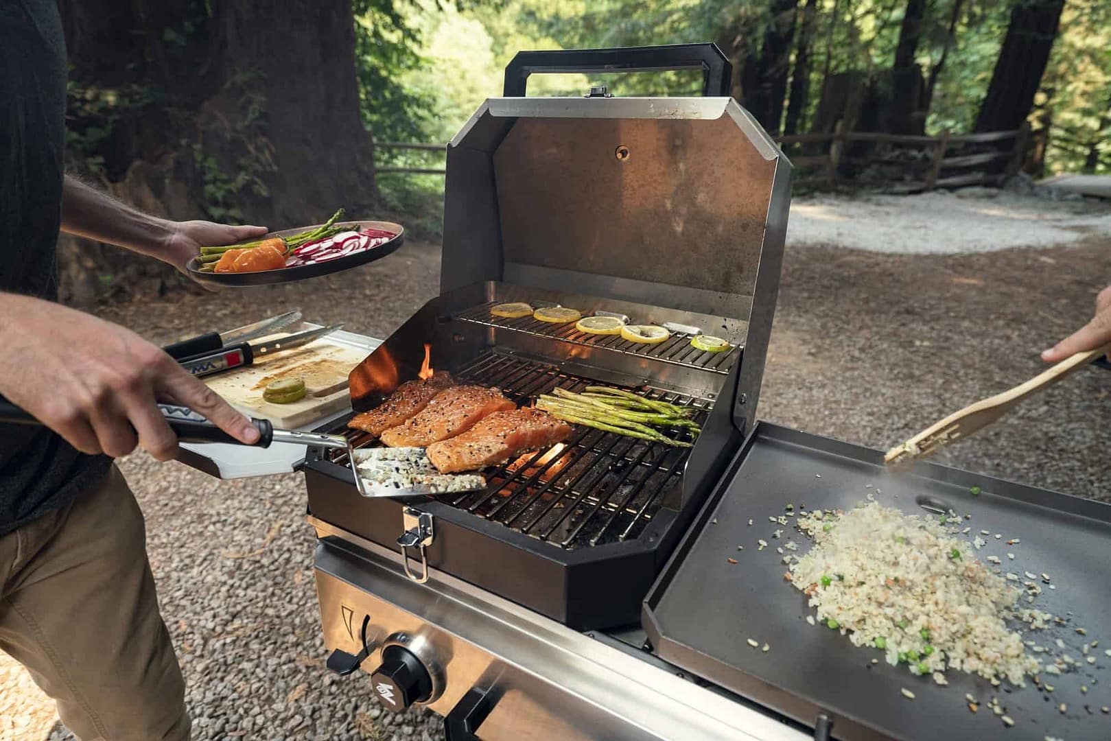 A man is grilling food on an outdoor grill in the woods.