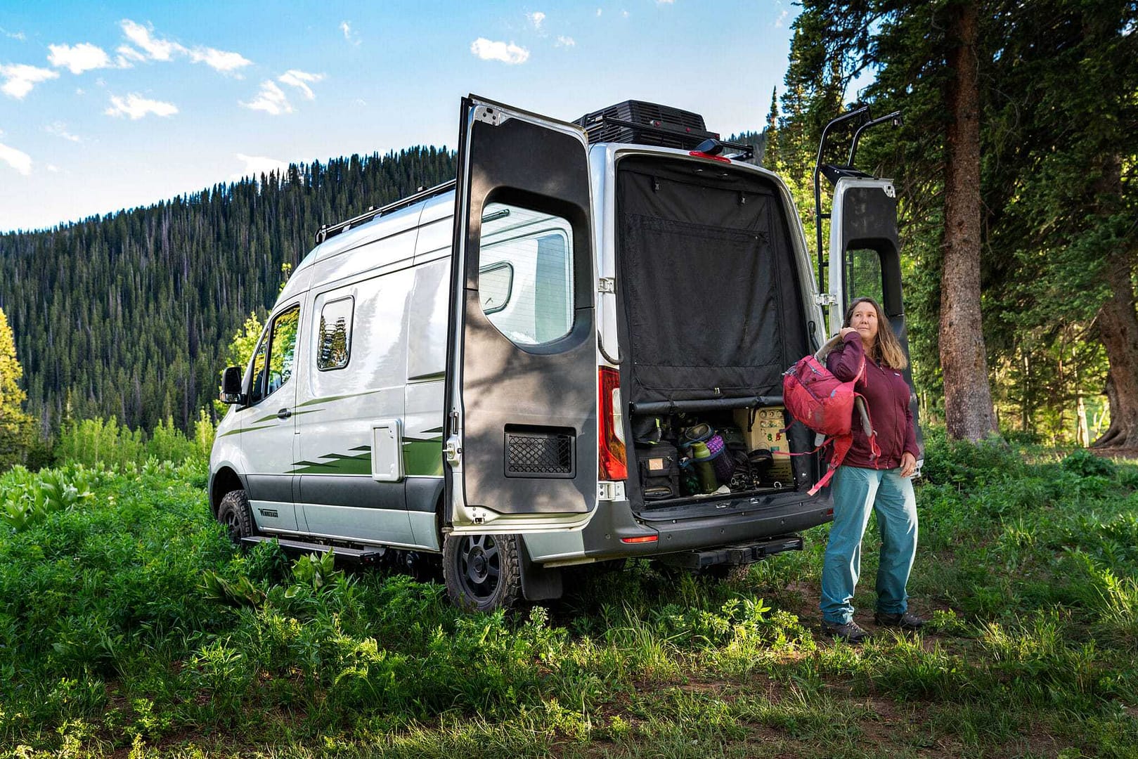 A woman standing next to a Winnebago camper van in the woods.