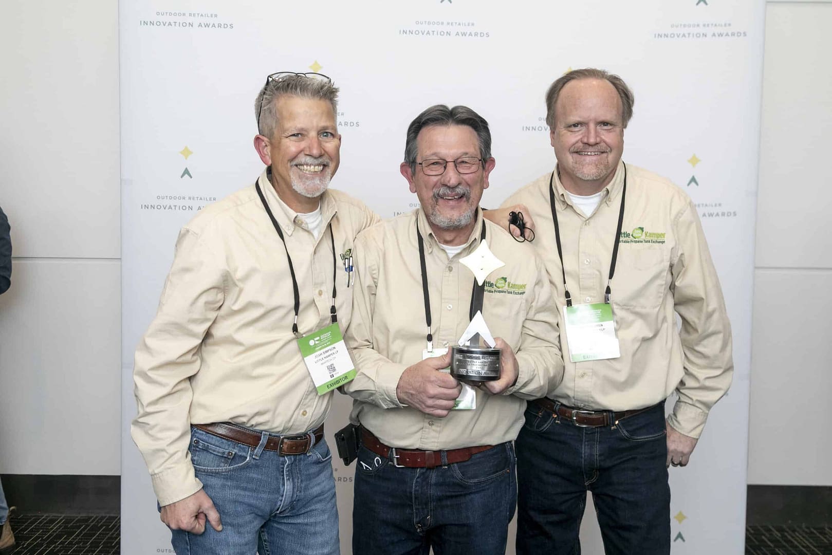 Three men posing for a picture in front of a banner at the awards ceremony for the Little Kamper Program.