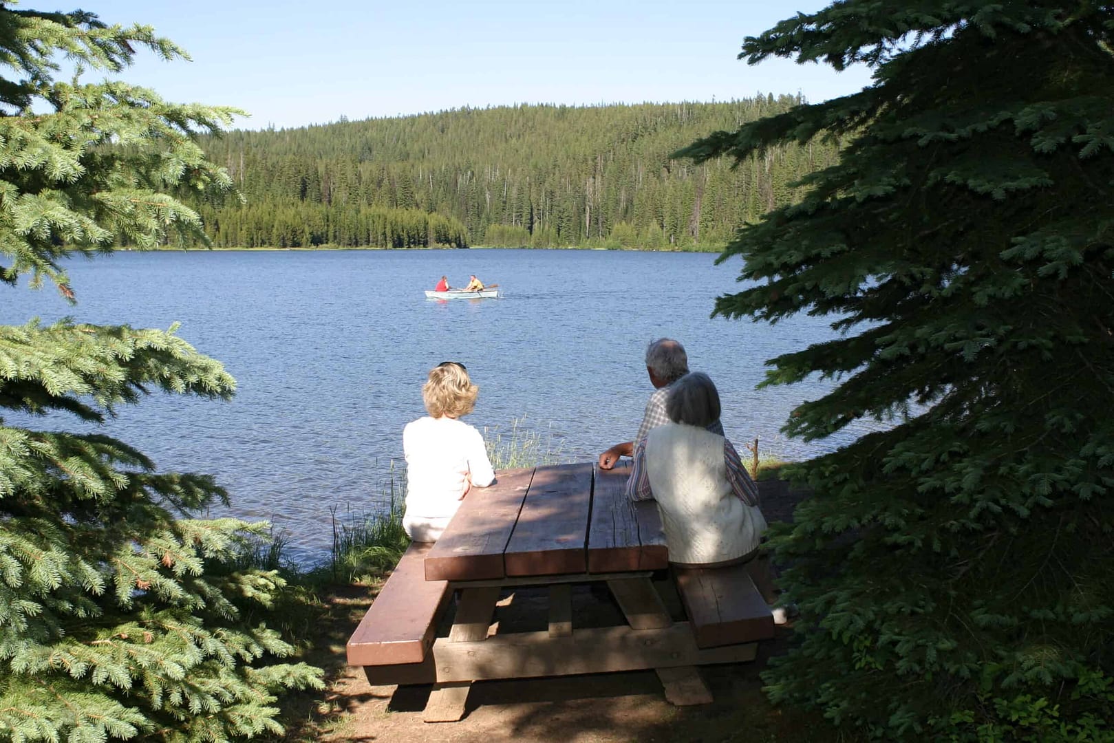 A group of Volunteer Campground Hosts sitting on a picnic bench in front of Umatilla National Forest lake.
