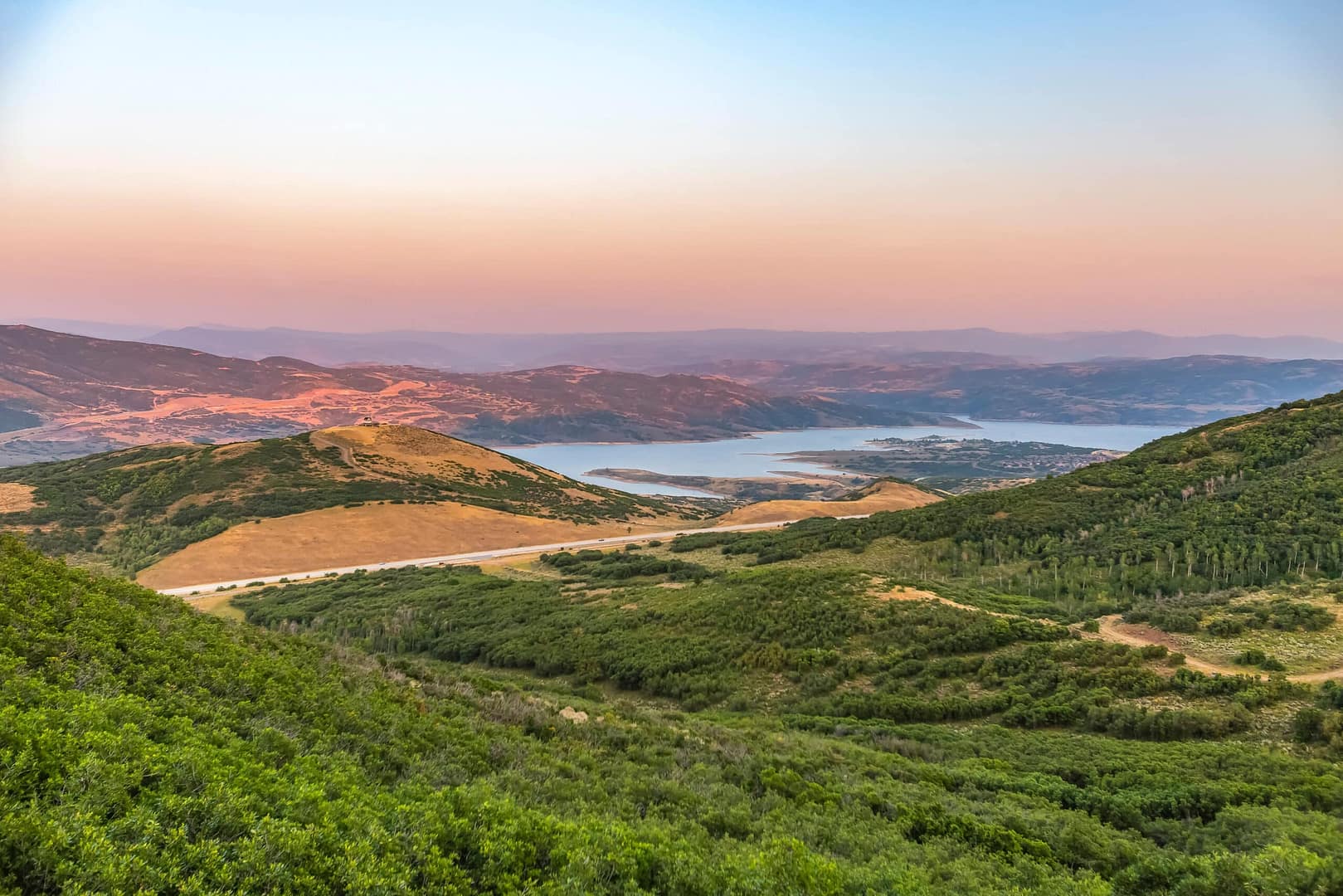 Sunset view over a hilly landscape with Jordanelle Reservoir and lush greenery, under a softly lit sky.