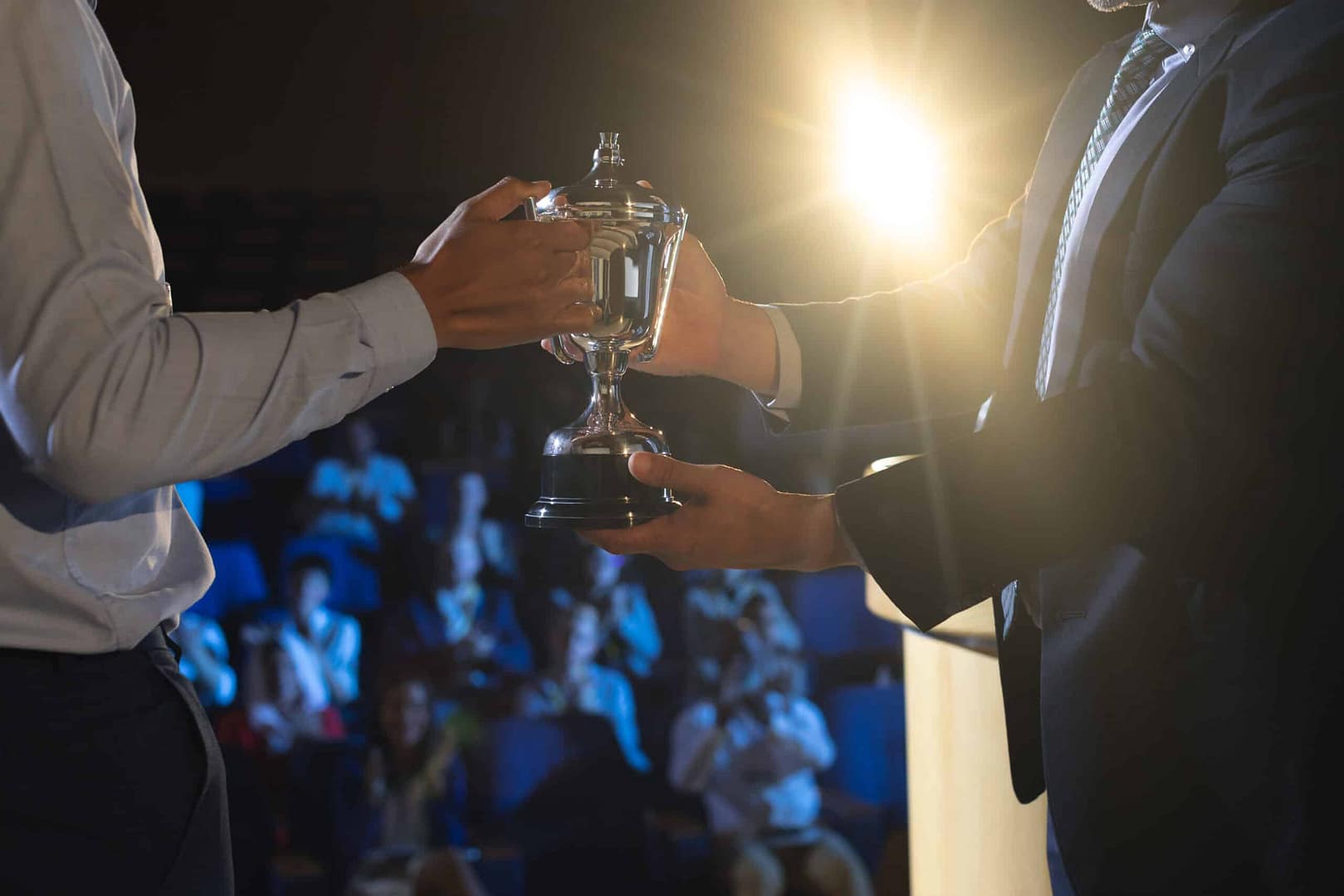Two people on a stage, handing a trophy to each other under a spotlight, with an audience in the background watching the auto draft event.