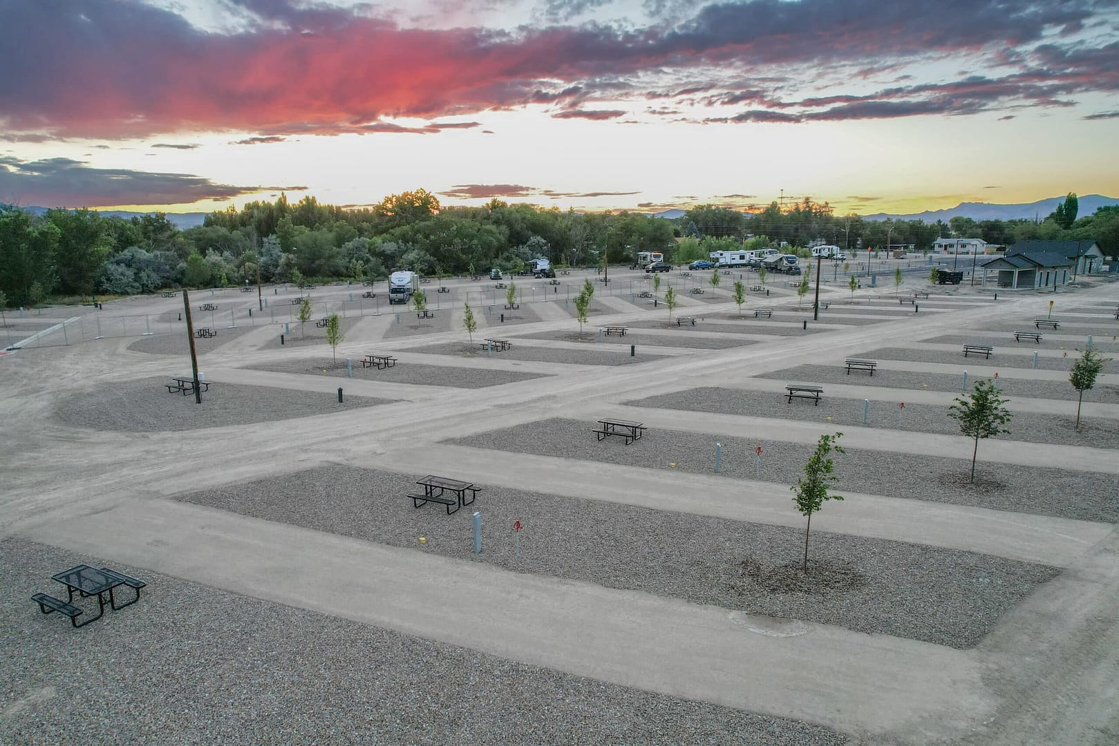 Aerial view of a campground at dusk with several RVs parked, picnic tables scattered around, and a colorful sky above.
