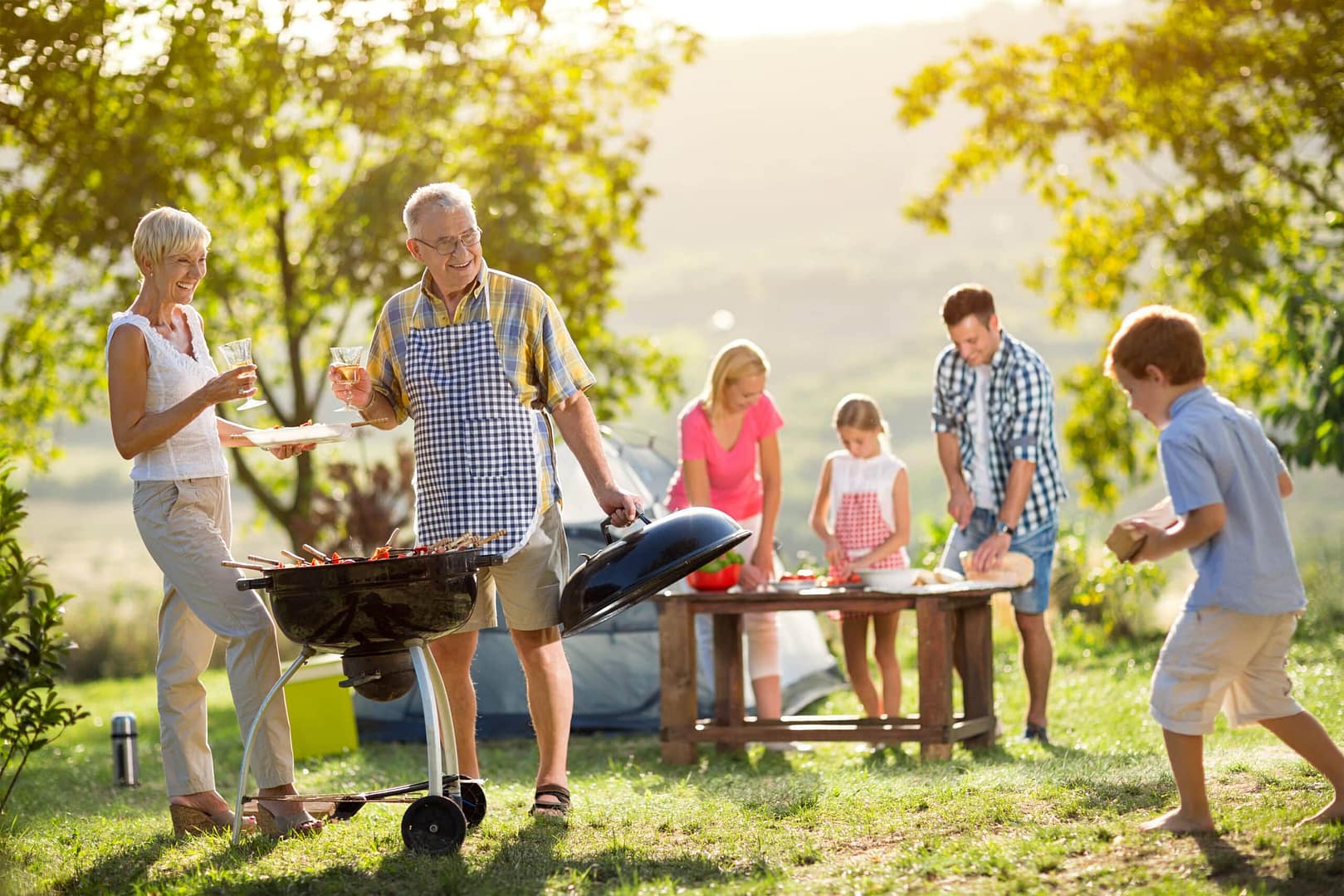 A family enjoys a barbecue in a sunny park, with an elderly man grilling, a woman eating, and others preparing food for their exploratory travel.