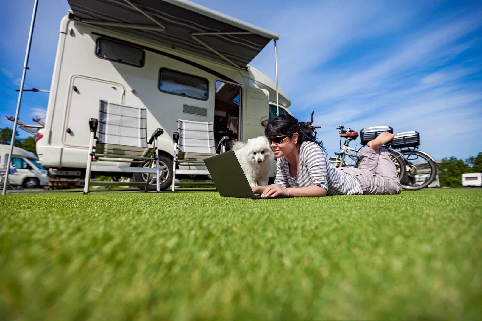 A woman lies on her stomach on the grass using a laptop next to her dog, with a camper van and bicycles in the background at an RV park.