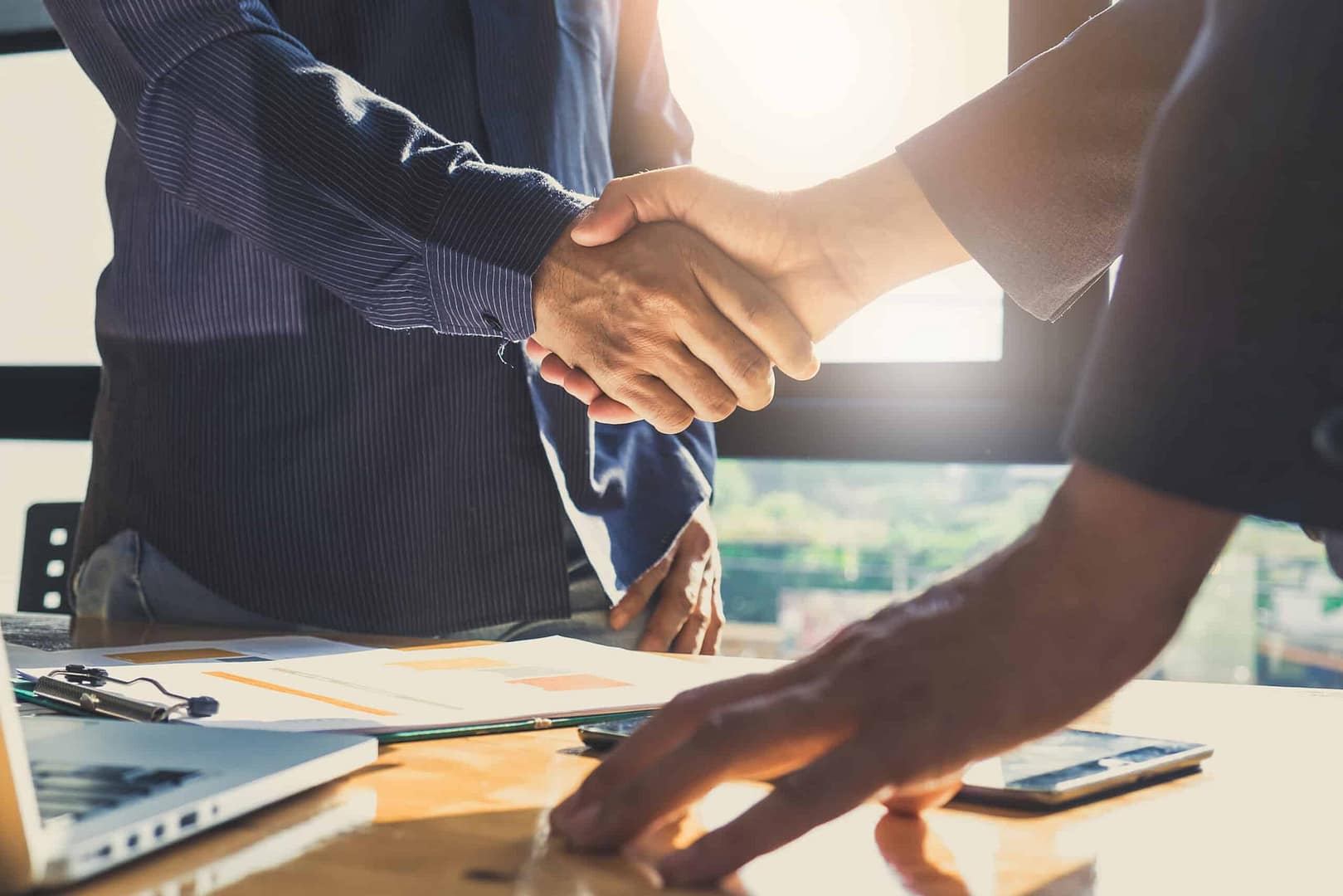 Two individuals shaking hands over a table with documents and digital devices, indicating an auto draft business agreement or partnership.