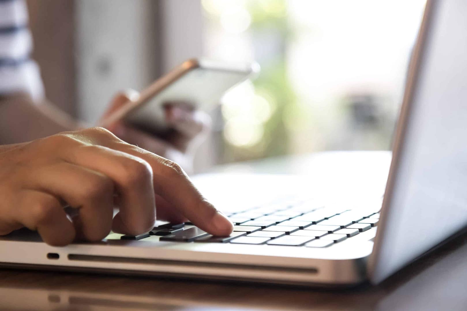 Close-up of a person's hand using a smartphone to review Campspot booking trends, with the other hand typing on a laptop keyboard, suggesting multitasking or digital interaction.