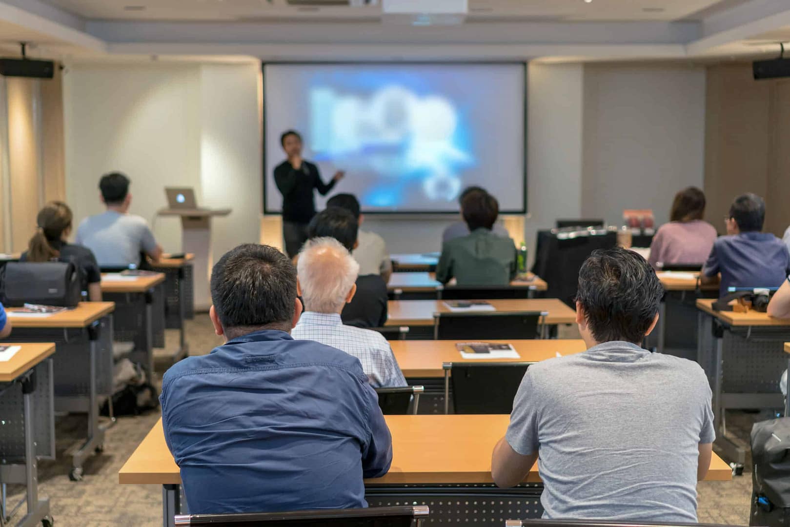 A group of people seated in a conference room, facing a presenter standing at the front near a screen displaying an auto draft of the presentation.