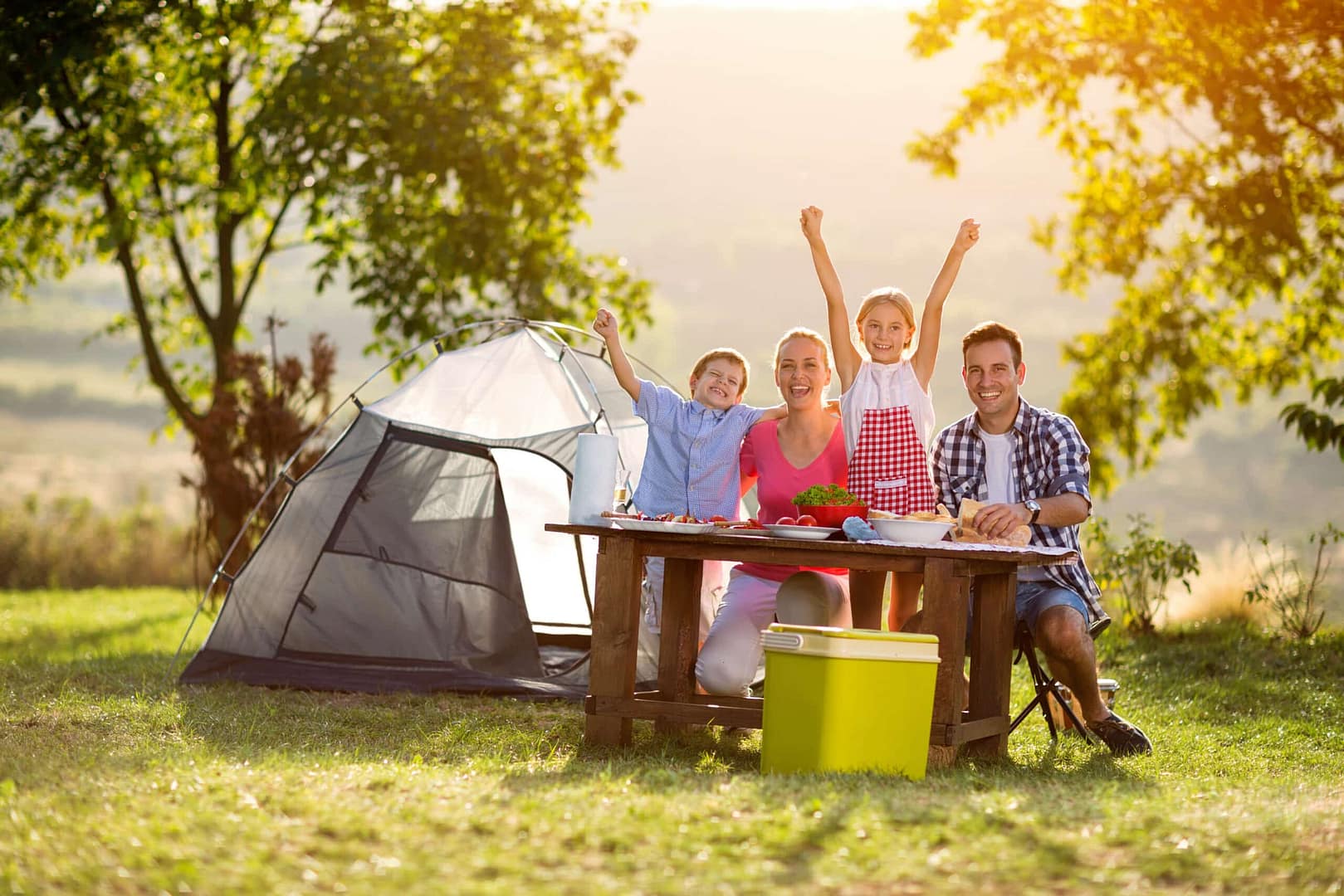 A family of four enjoying a picnic next to a tent in Schafer State Park, on a sunny, grassy field with one child playfully raising her arms in joy.
