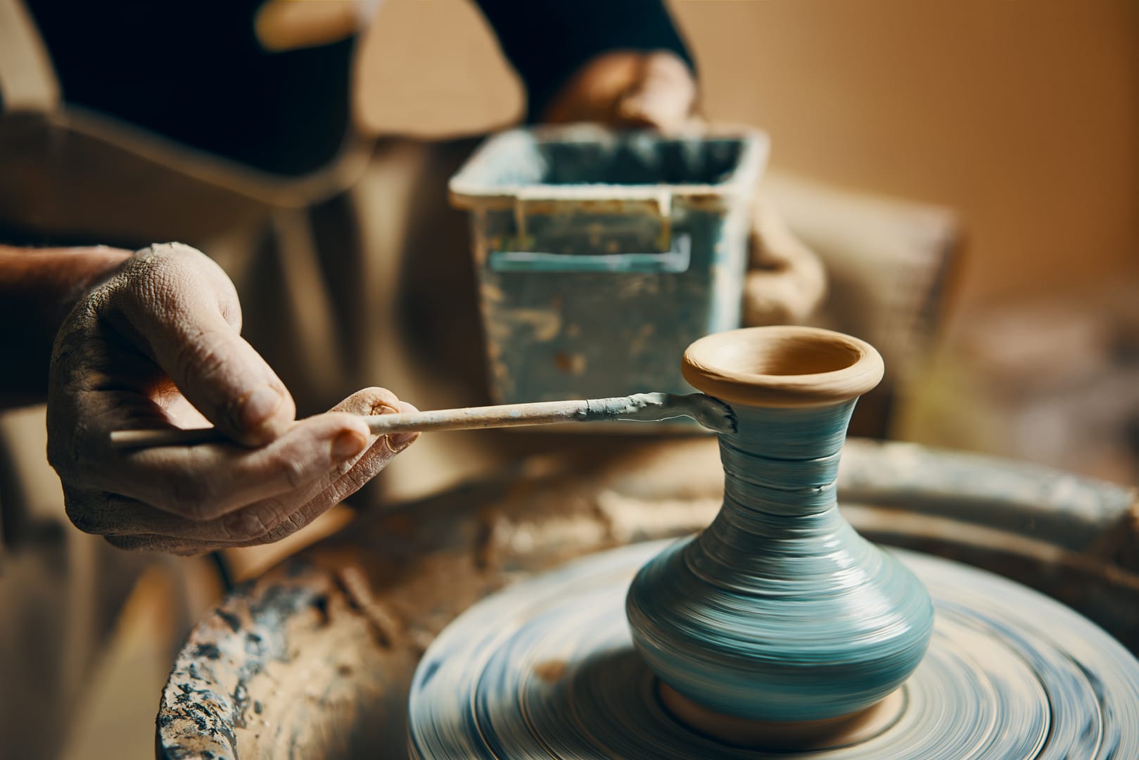 A guest immerses themselves in the experience by applying glaze to a partially finished blue pottery vase on a spinning pottery wheel, as part of our arts and crafts programs.