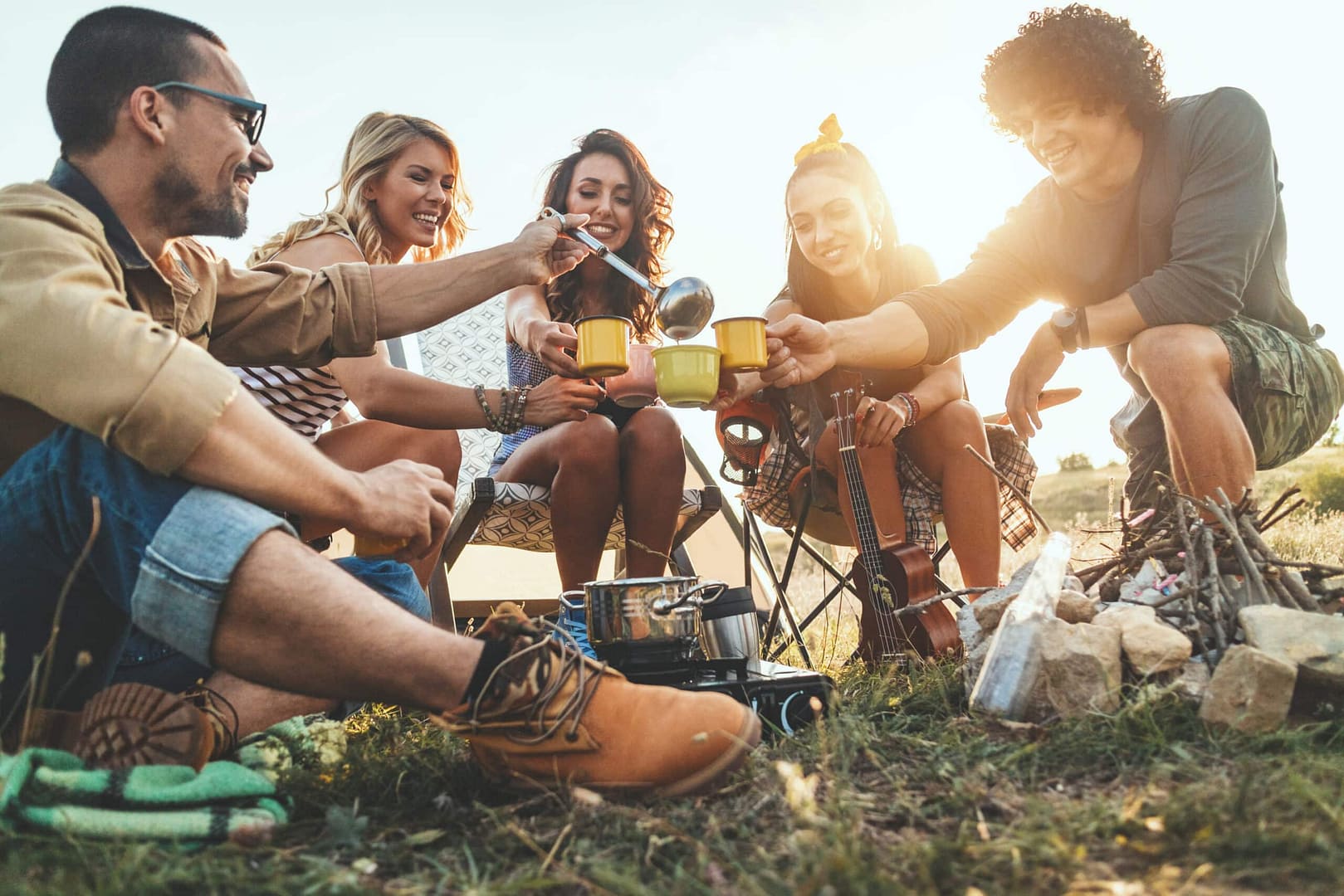 A group of young adults sitting outdoors, laughing and sharing food around a small campfire at Starved Rock Campground during sunset.