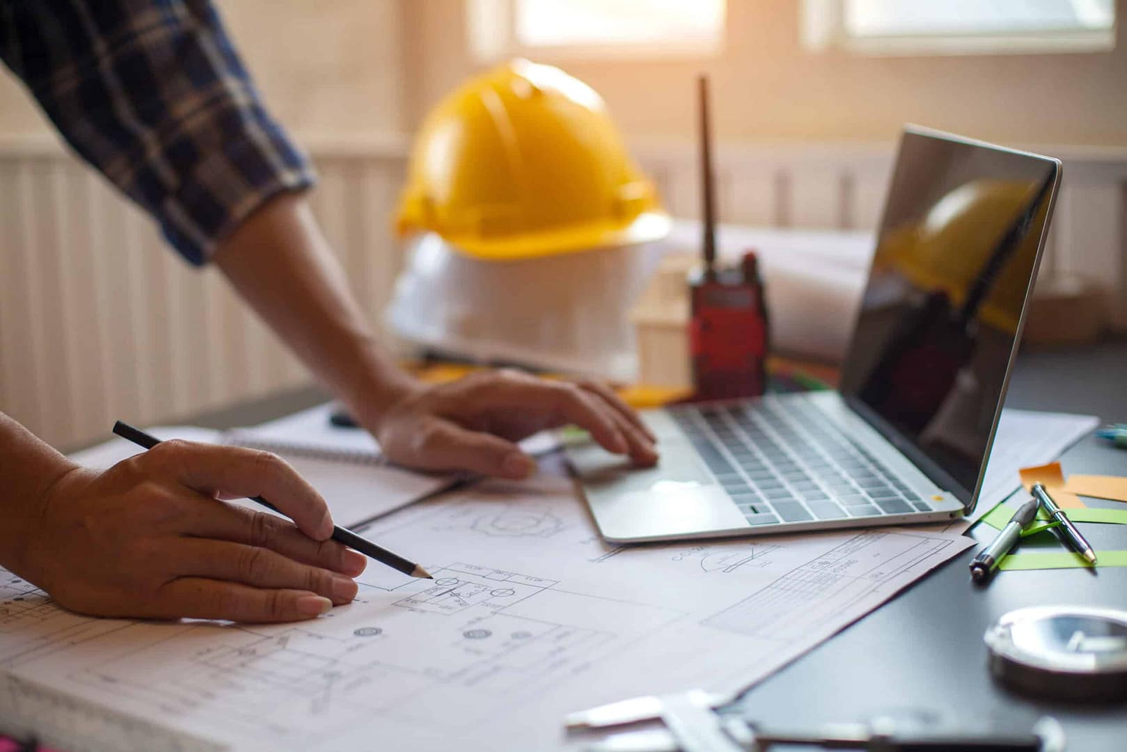 Person working on architectural plans with a pencil next to a laptop on a desk, utilizing Auto Draft for precision, with a yellow hardhat and a walkie-talkie in the background.