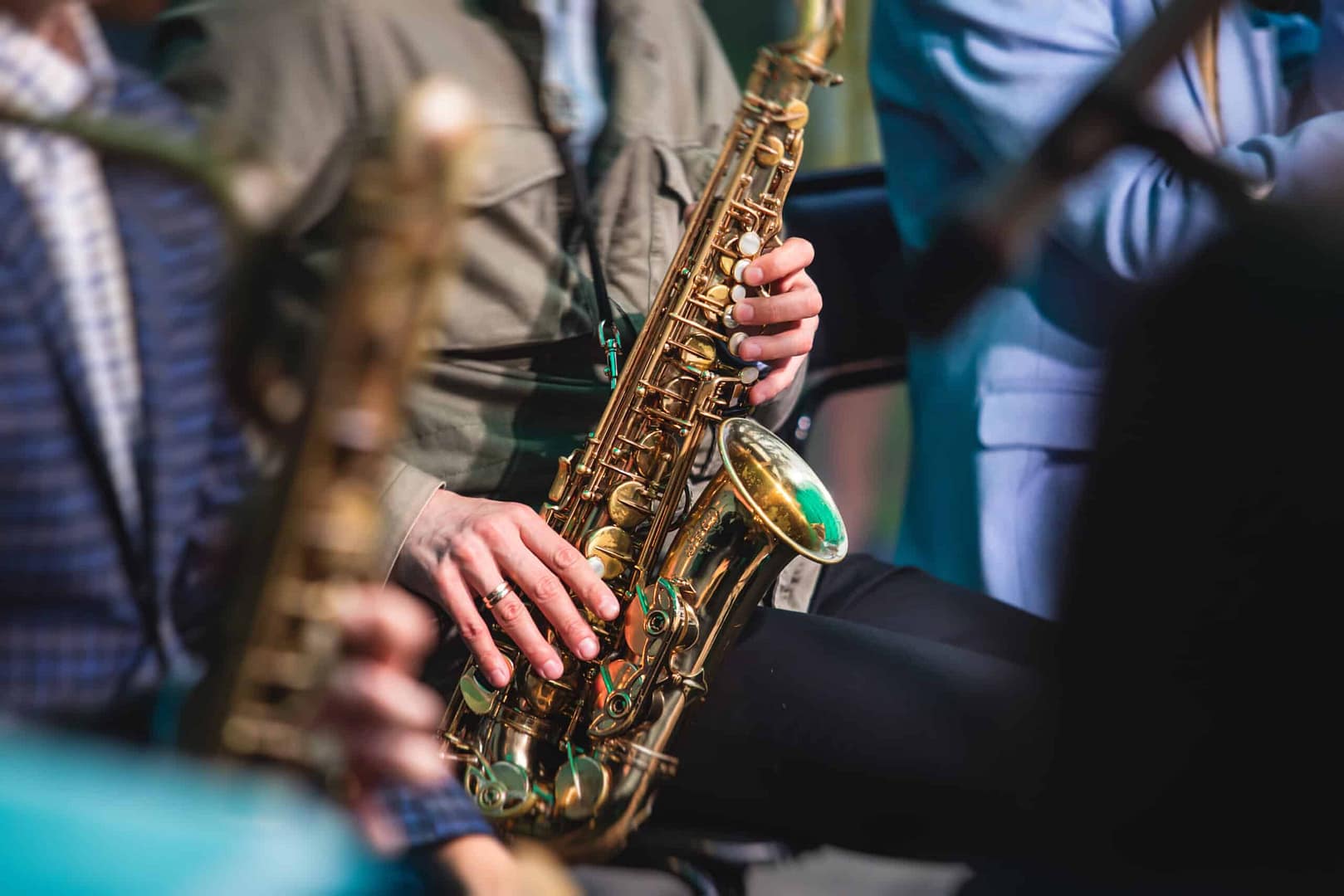 A person playing a saxophone with a close-up on the instrument and their hands, surrounded by other musicians. The setting appears to be a live performance or rehearsal, celebrating local music and engaging guests with vibrant performances.