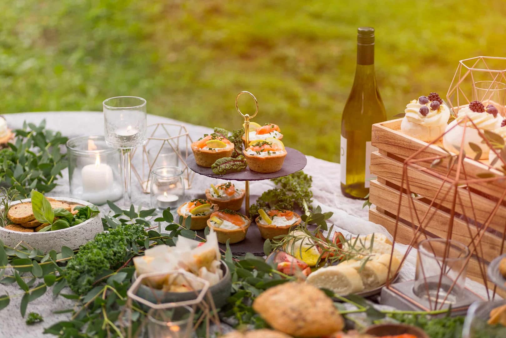 A spread of various appetizers, bread, and desserts on an outdoor picnic table, with a bottle of wine, decorative candles, and greenery embellishments highlights the farm-to-table dining experience for guests.