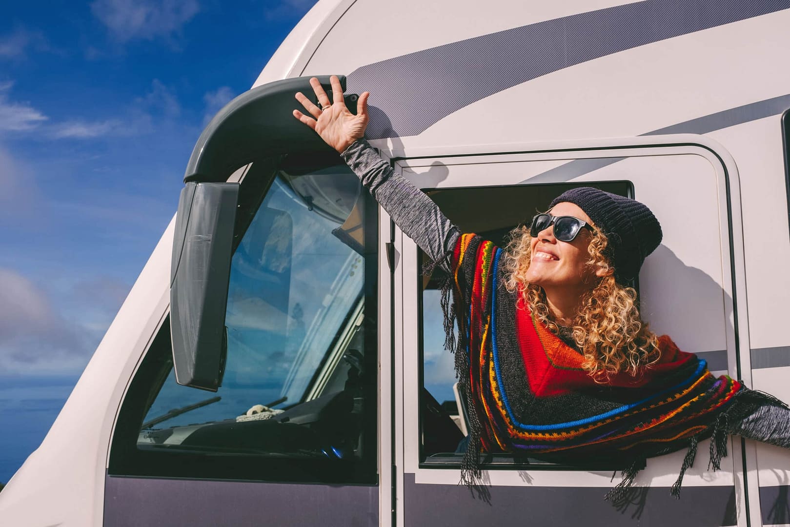 Woman in sunglasses and scarf smiling and stretching beside an auto draft camper van under a clear blue sky.