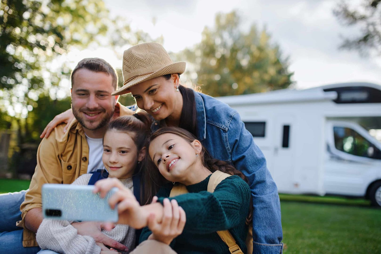 A family of four, two adults and two children, take a selfie together outside near a white RV, with trees in the background—capturing their spontaneous adventure in an auto draft of memories.