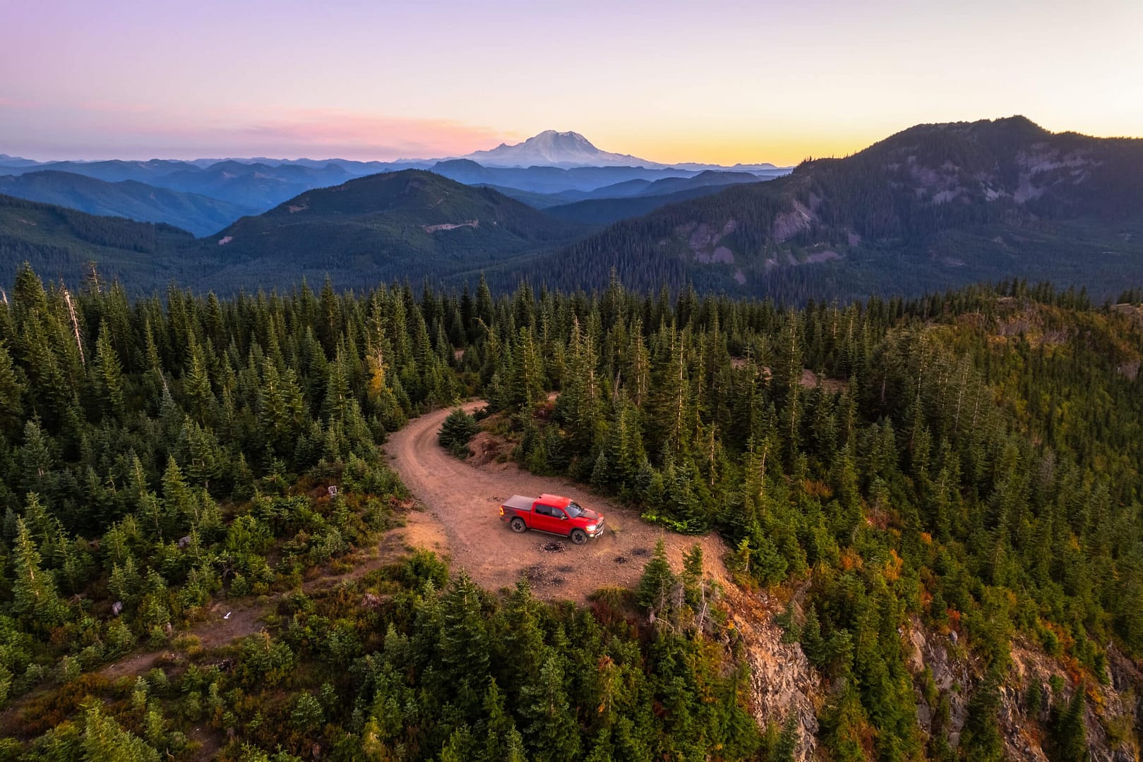 Aerial view of a red SUV, now a camping vehicle, parked on a winding dirt road in a dense forest at sunset, with mountains in the background.