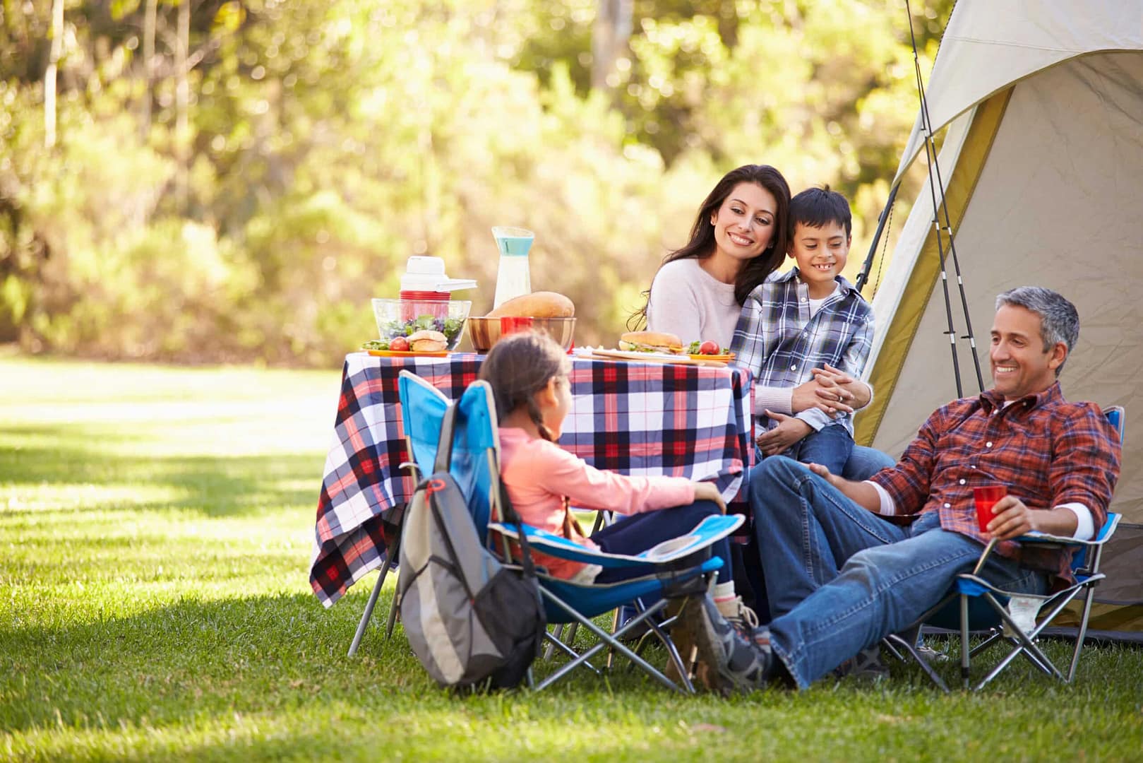 A family of four enjoying an inclusive camping picnic near a tent in a sunny park, with a plaid-covered table full of food.