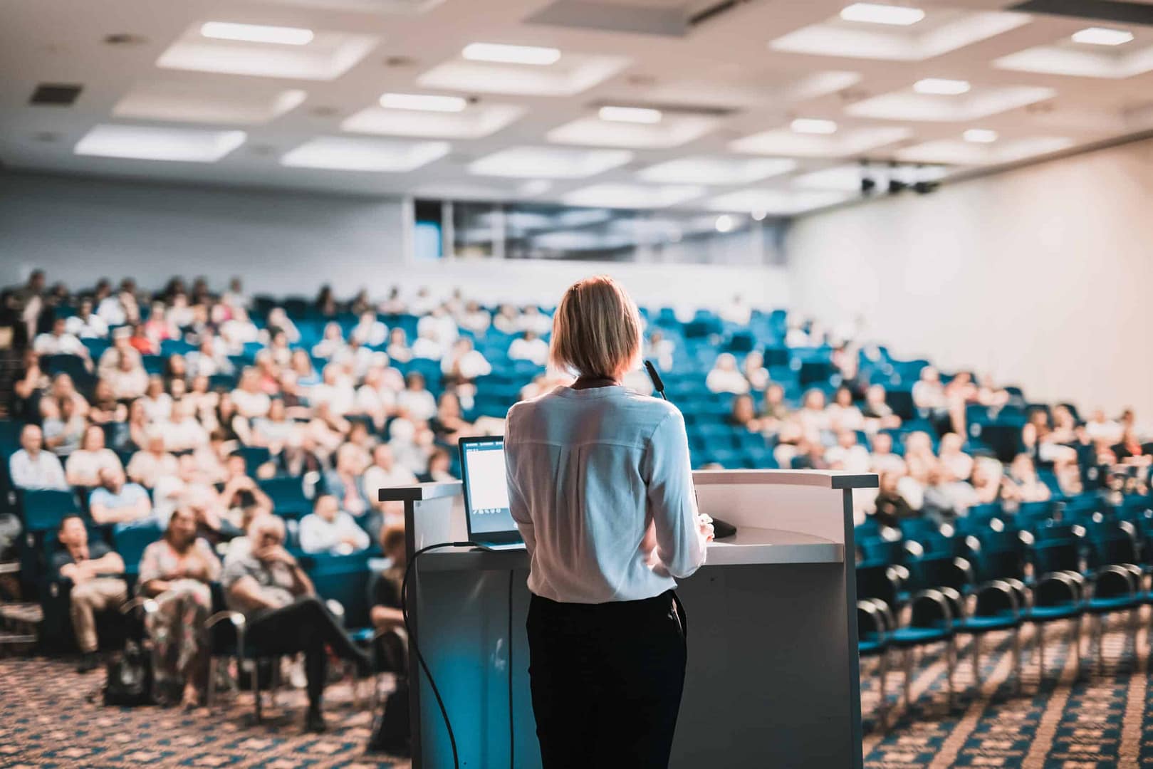 A woman presenting at a podium in a conference room filled with attendees from the camping industry.