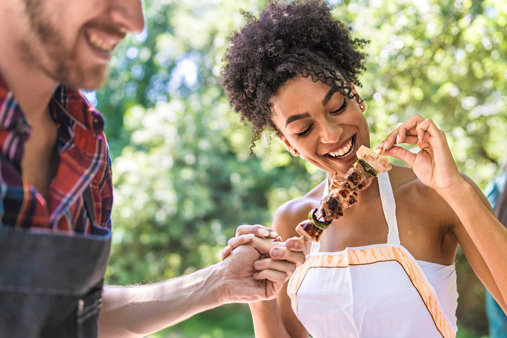 At a vibrant campground, a woman in a white top smiles while enjoying a skewer of grilled meat and vegetables. A man in a plaid shirt and apron holds her hand, both embracing the local culinary experiences outdoors.