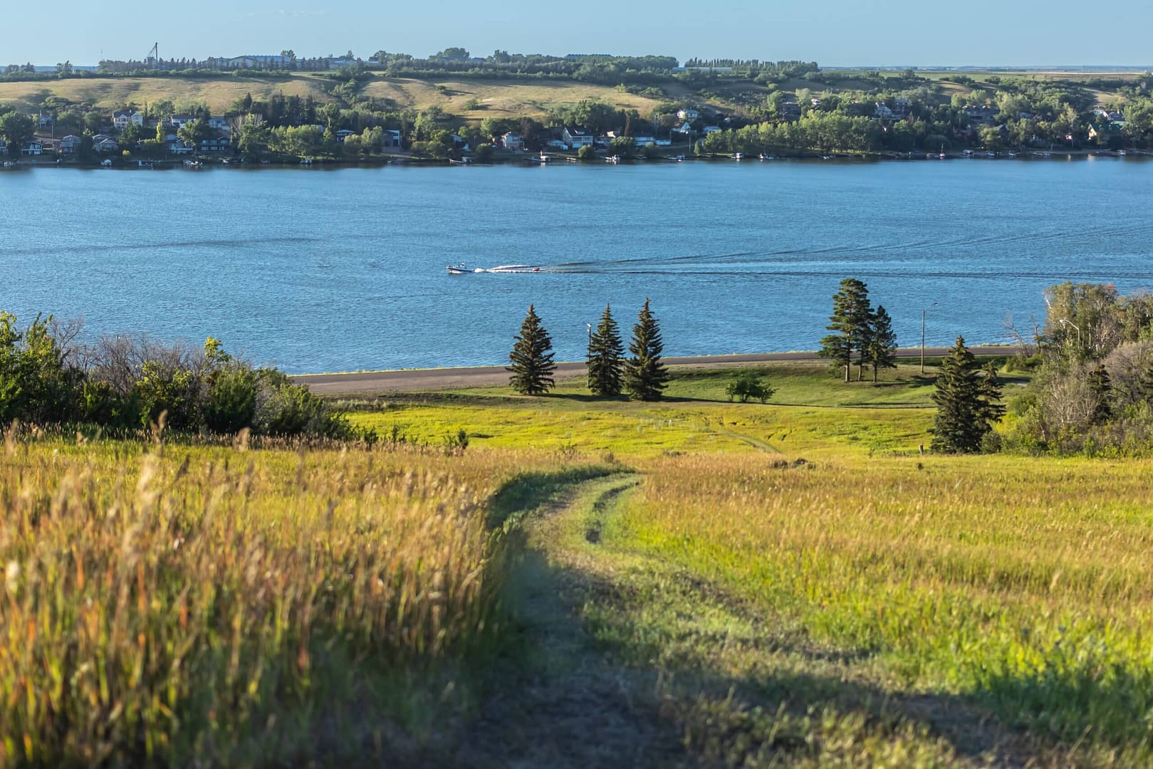 A dirt path leads through a grassy field toward a lake in Saskatchewan Provincial Parks, with a few trees near the shore. A boat creates a wake on the lake, and houses are visible on the distant shoreline. As camping season approaches, new features and programs await adventurers.