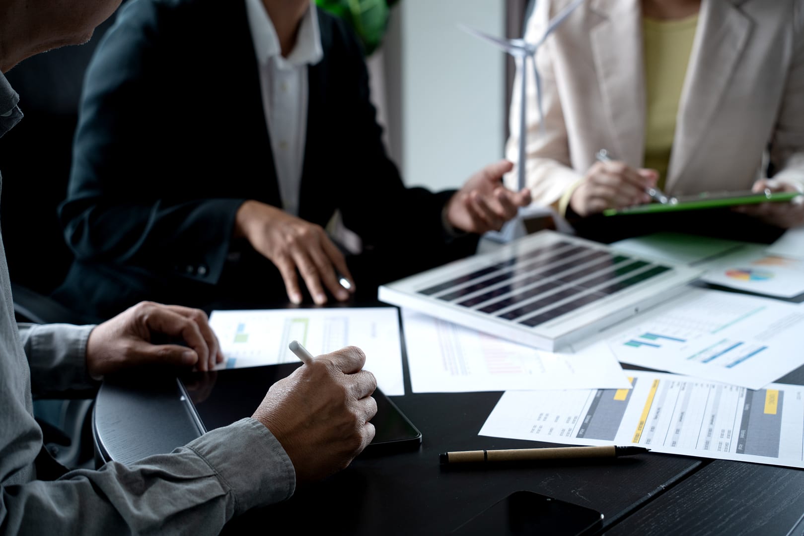 Professionals discussing renewable energy projects with wind turbine model and solar panels on an auto draft table.