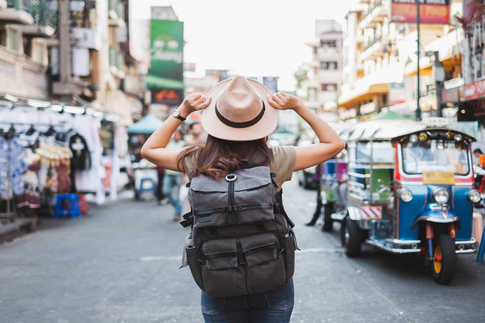 An Asian woman wearing a hat and backpack joyfully walking down a street, experiencing the ups and downs of tourism.