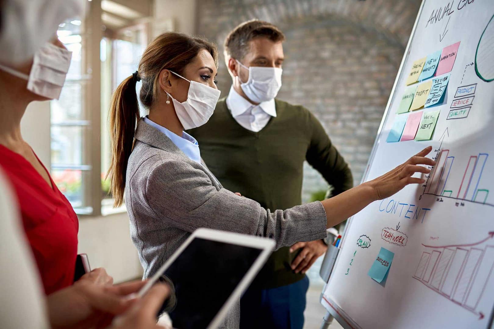 A proposed group of people wearing face masks are working on a whiteboard, discussing changes.