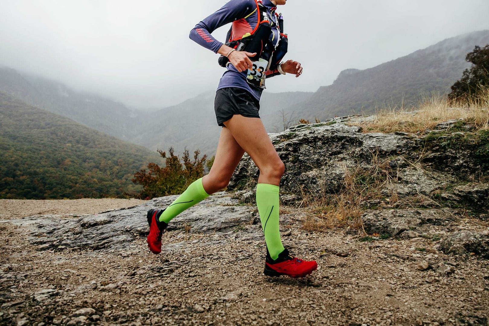 A woman wearing an Ultimate Direction Ultralight Vest is running on a mountain trail.