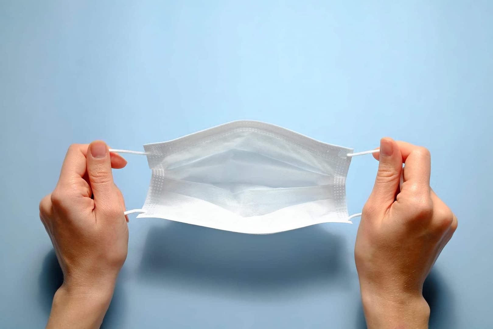 A pair of hands holding a disposable surgical mask on a blue background in New Zealand.
