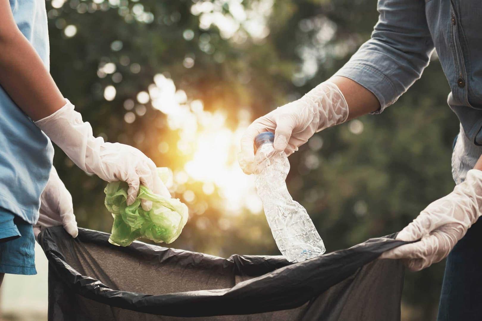 Two people in gloves are putting trash into a trash can.