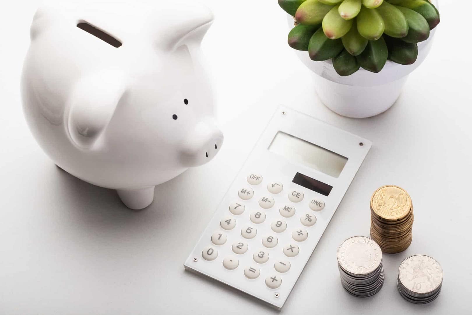 A piggy bank filled with coins sits next to a plant on a desk, while a calculator is used for digital financing calculations.