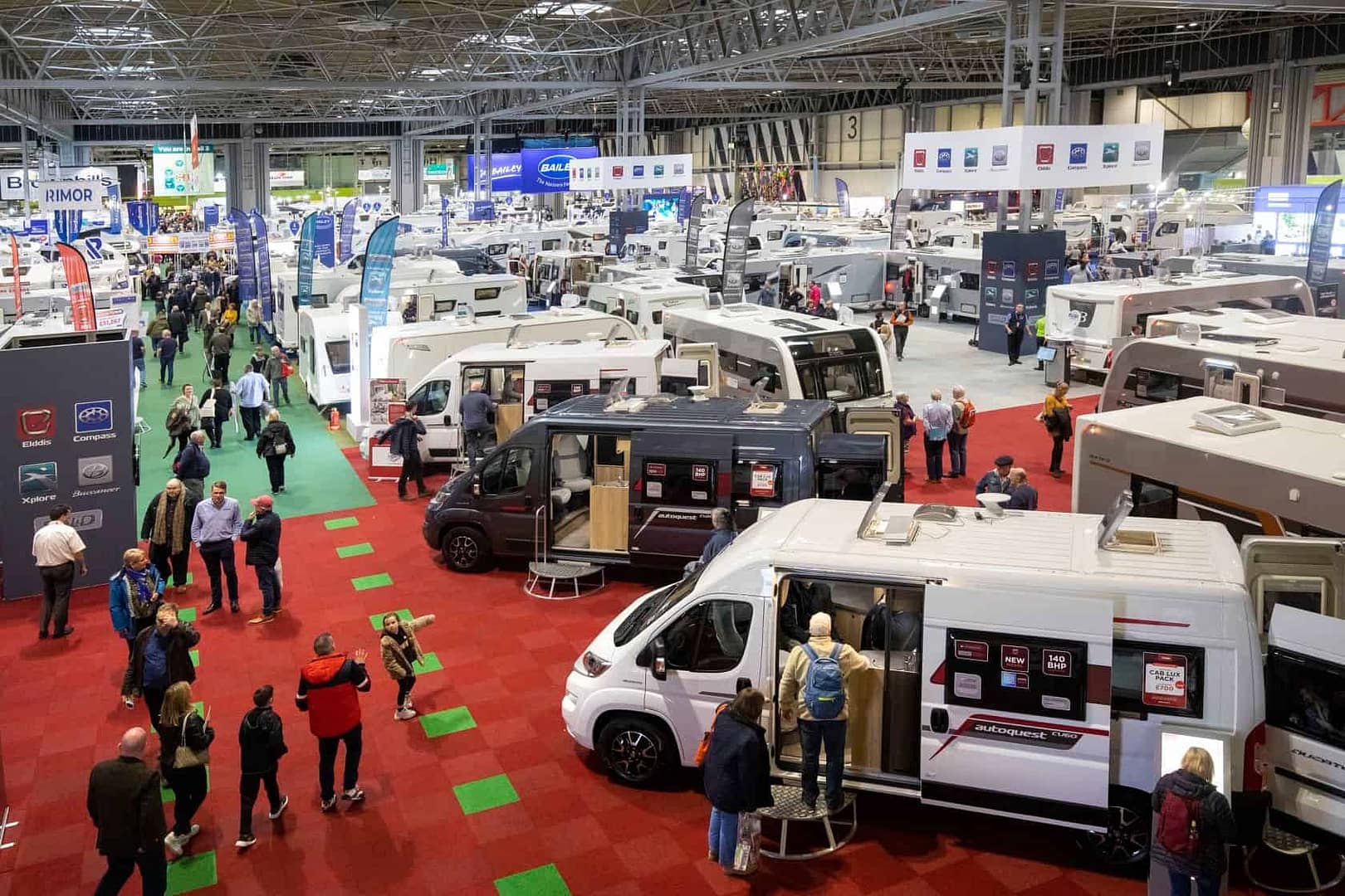 Attendees at the UK Caravan Camping & Motorhome Show gather in a room filled with many RVs, eagerly anticipating the Speaker Lineup presentations.
