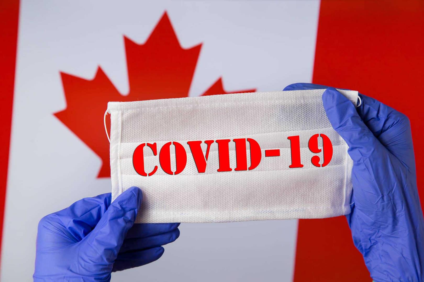 A person holding a covid-19 mask in front of a Canadian flag with Manitoba as the backdrop.