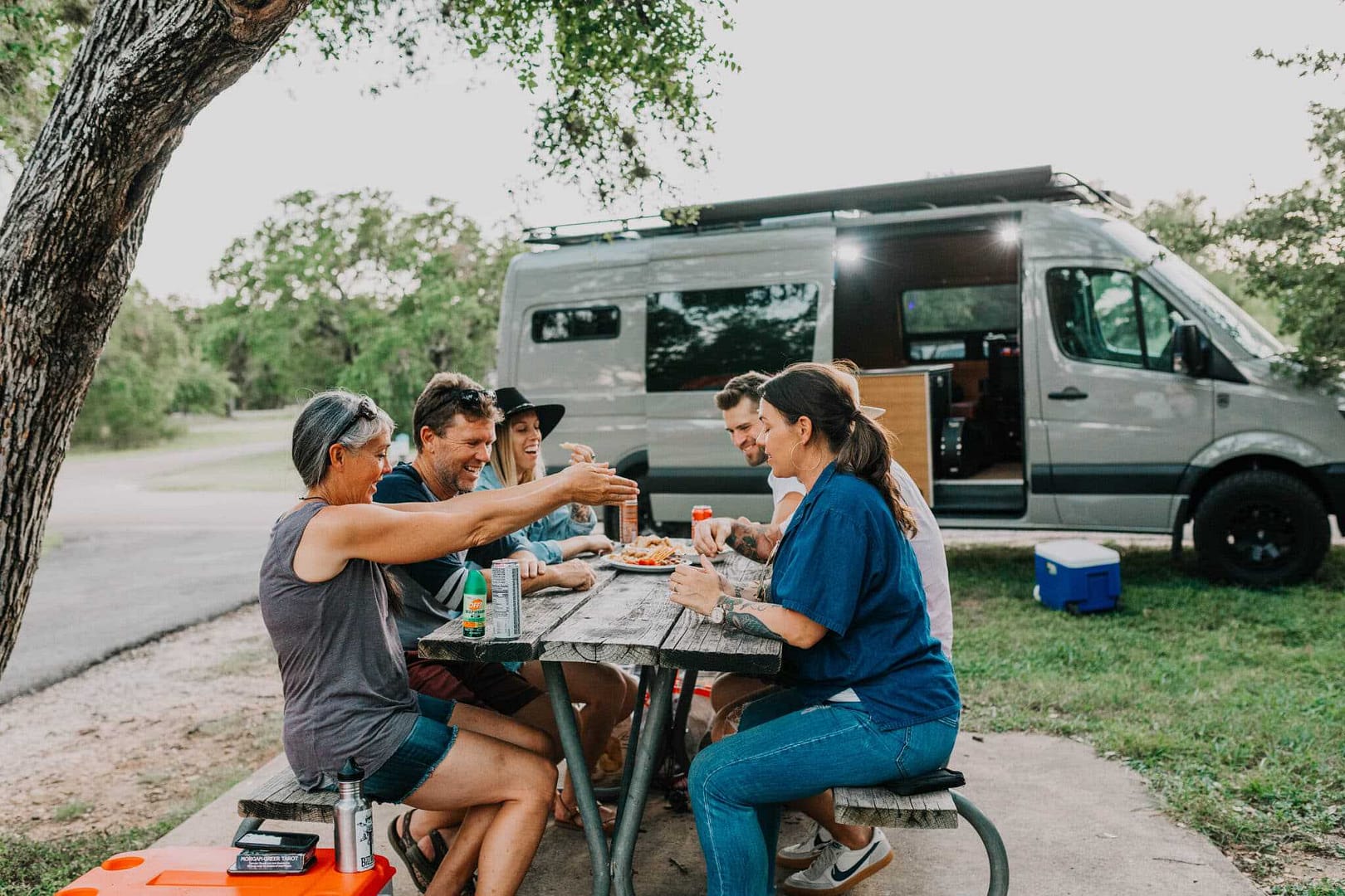 A group of people sitting at a picnic table near an rv. The group consists of employees from Forbes and Outdoorsy, two notable startup employers.