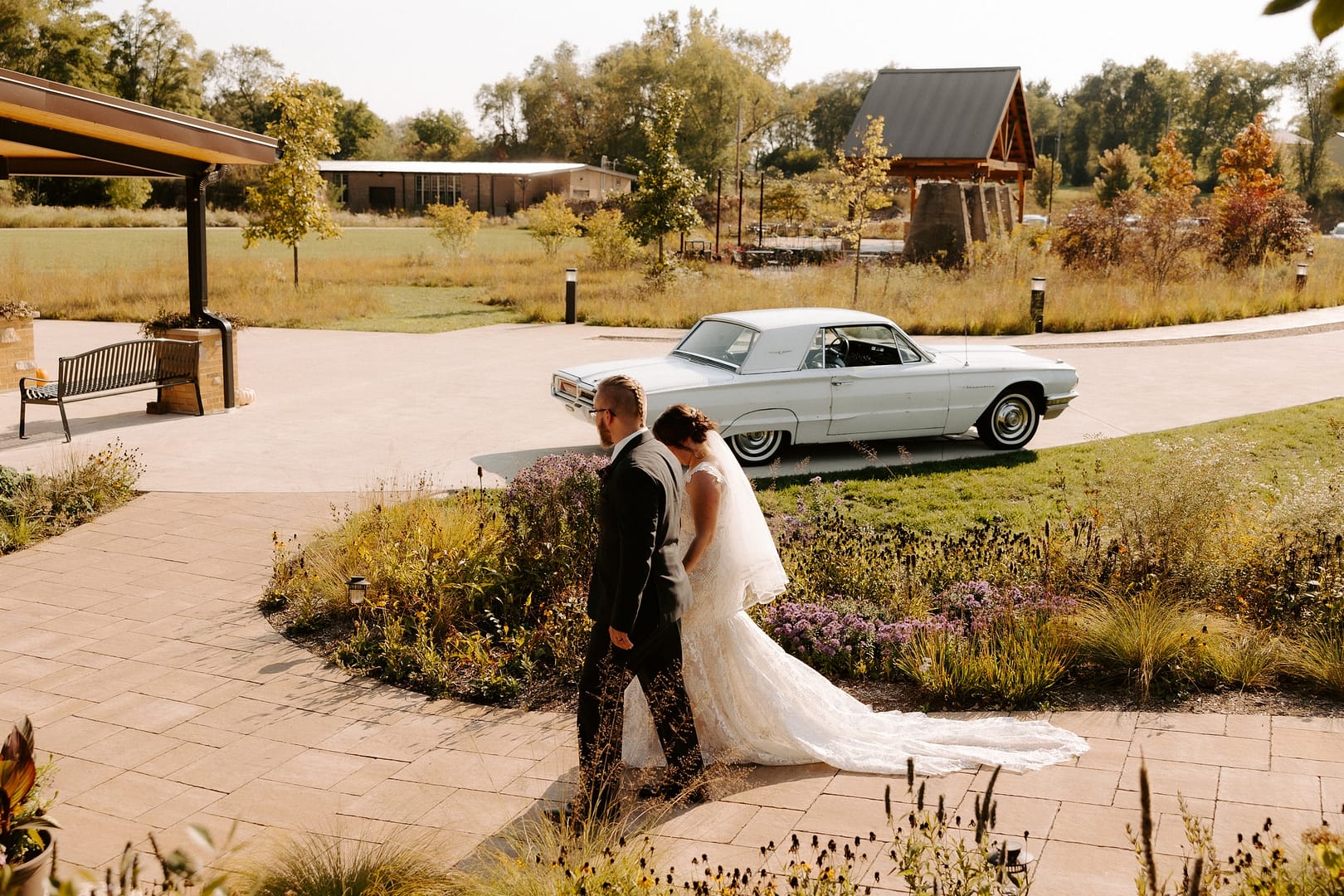 A couple celebrating their wedding day poses in front of a classic car at a picturesque park.