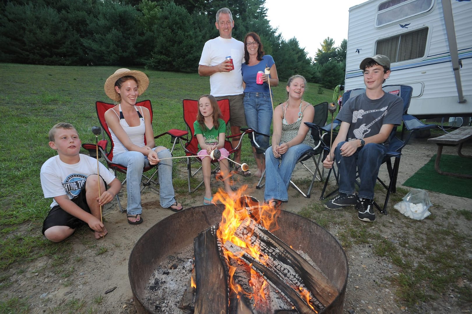 A family of outdoor lovers sits around a campfire in front of an RV at Lake George RV Park.