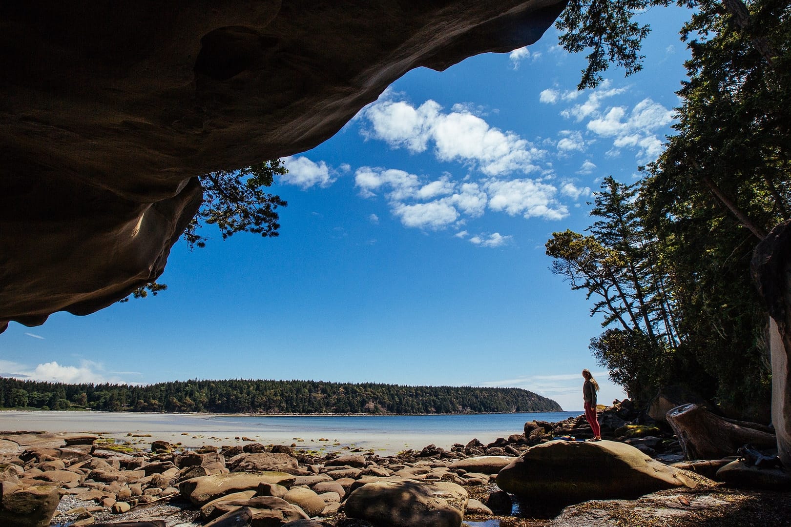 A person stands under a large rock overhang, looking at a scenic view of a rocky beach and tree-lined coast under a blue sky.
