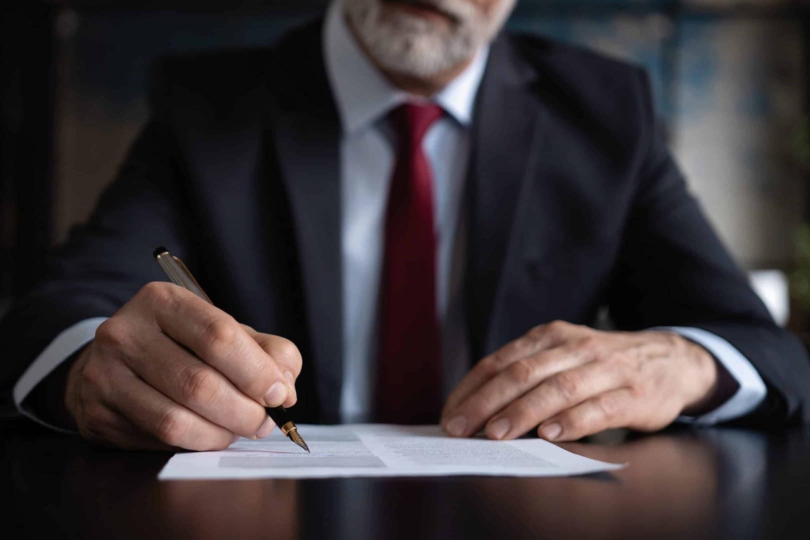 A senior businessman in a suit and red tie signing a USDA document at a desk.